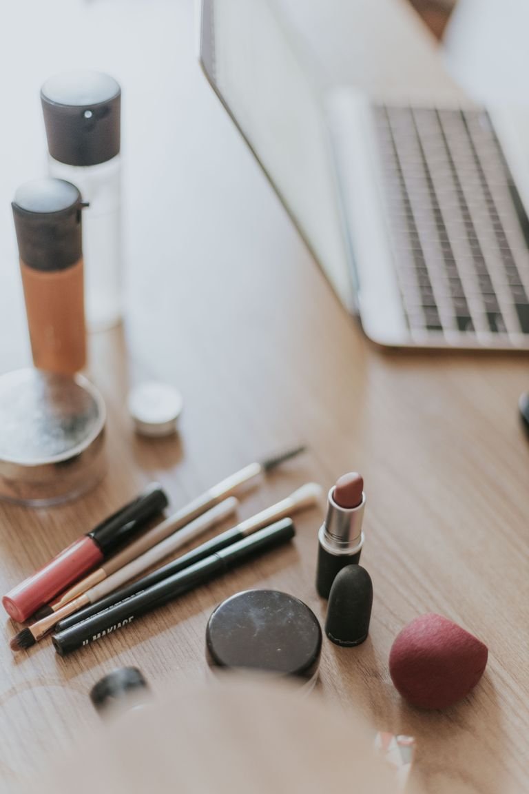 A collection of makeup items alongside a laptop on a wooden desk, ideal for beauty work settings.