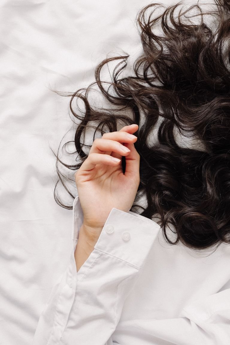 A delicate hand in a white shirt touching wavy dark hair on a bed.