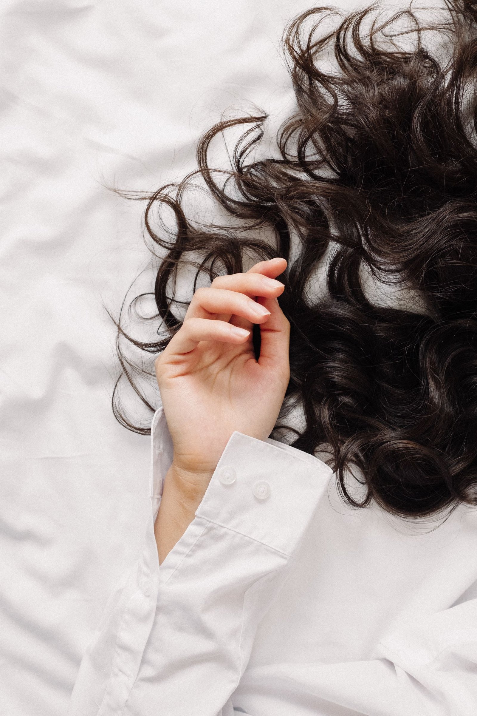 A delicate hand in a white shirt touching wavy dark hair on a bed.
