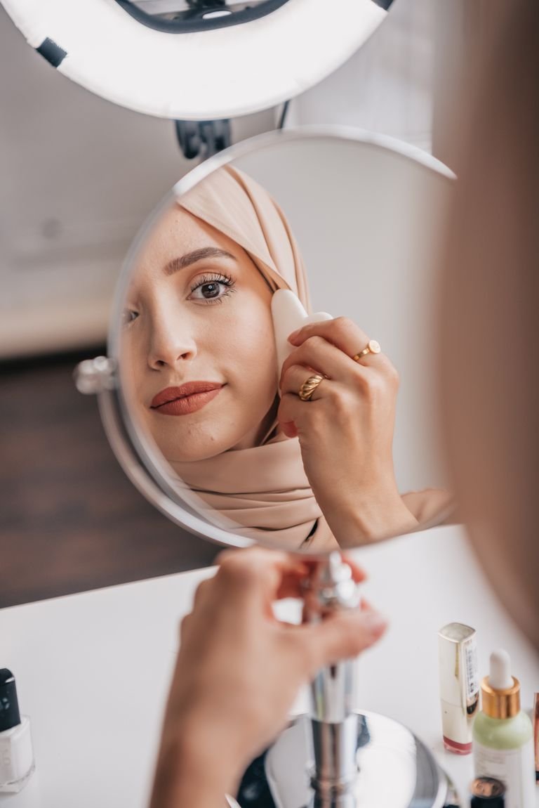 Middle Eastern woman in hijab using a gua sha tool in front of a mirror for a beauty routine.