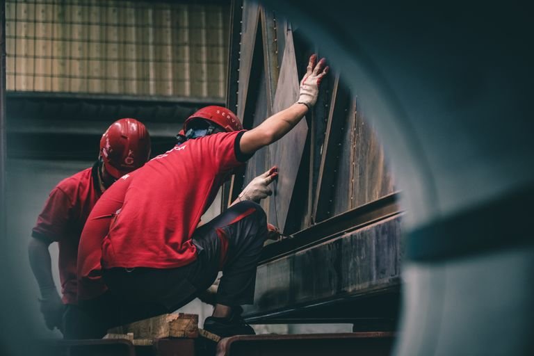 Two workers in red helmets assembling metal components inside a factory.